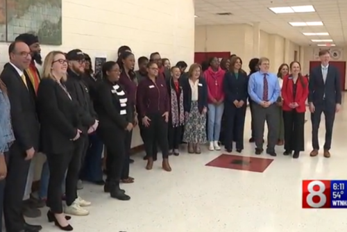 25 city leaders stand in the hallway of Wilbur Cross High School.