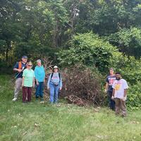 Volunteers stand with a pile of dead invasive plants that they have removed from Fairmont Park. The image shows seven people and they are on a field at the edge of a forest.