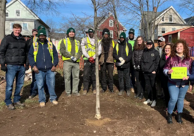 Tree planting crew and volunteers stand behind the new tree. A woman holds a paper sign saying "Tree #1."