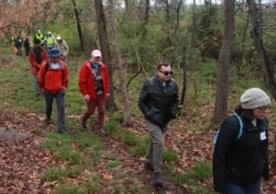 10 adults walk through the wooded trail dressed for a cool spring day.